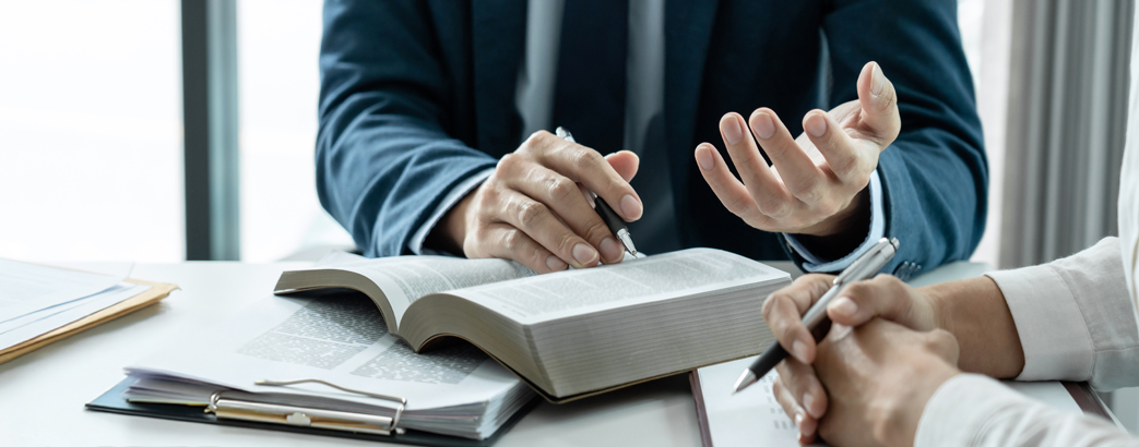Two people in formal attire sit at a table with legal books and documents. One person's hands are gesturing while the other's are holding a pen, as if discussing or consulting something legal. A gavel