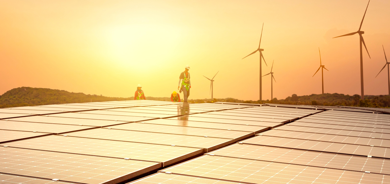 Workers in safety gear inspect solar panels on a rooftop at sunset. Wind turbines are visible in the background, set against a hilly landscape. The scene highlights renewable energy sources with a war