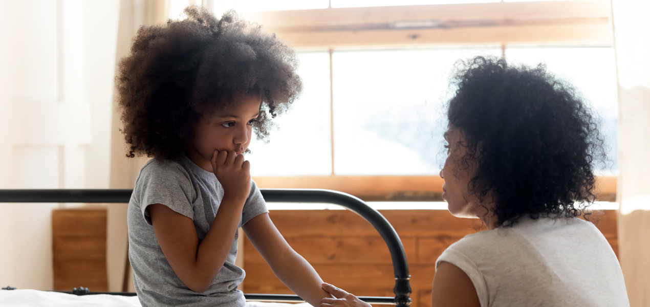 A woman kneels beside a bed, holding the hands of a young girl who is sitting on it. The girl appears thoughtful or concerned.