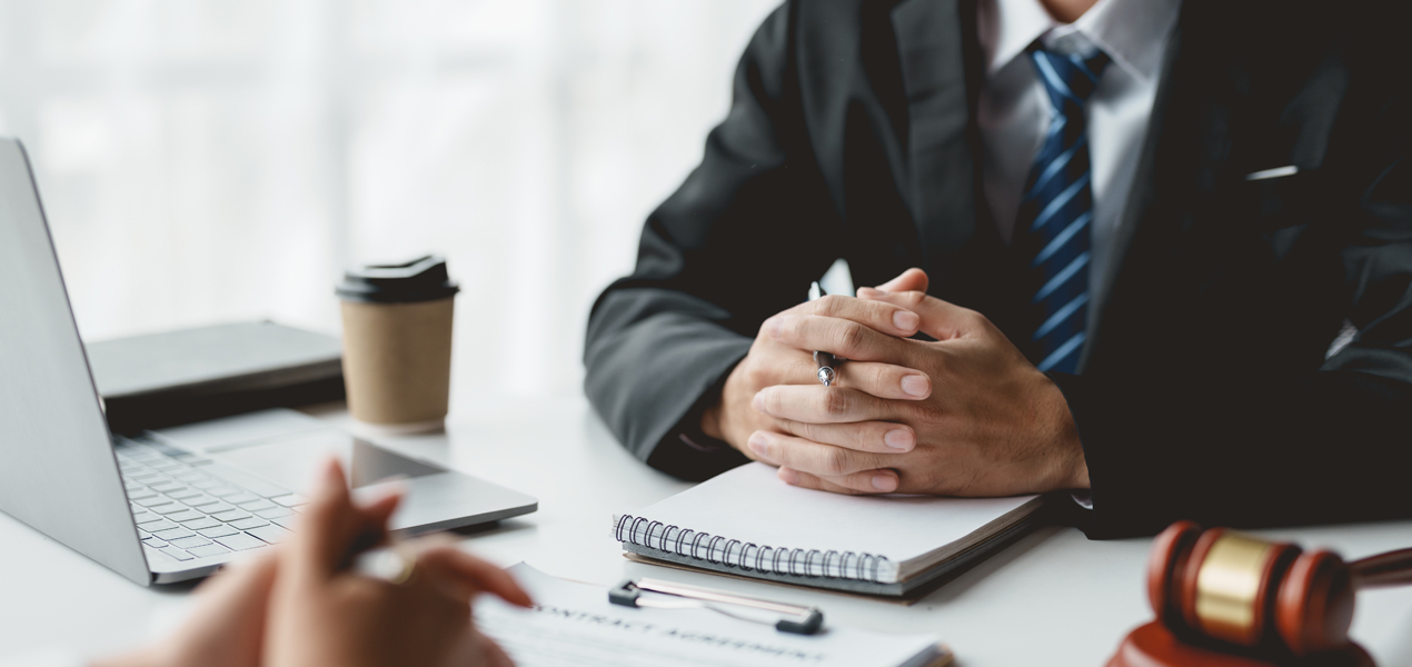 Two people sitting at a desk with papers, a laptop, and a gavel. One person in a suit has hands clasped. The focus is on the business setting, suggesting a formal or legal discussion.