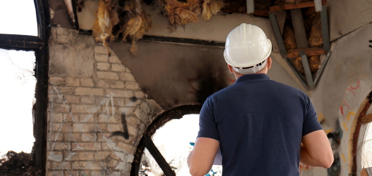 A person in a hard hat and blue shirt is inspecting a damaged building interior. The walls are charred, and insulation is exposed from the ceiling. Broken windows and debris suggest significant struct